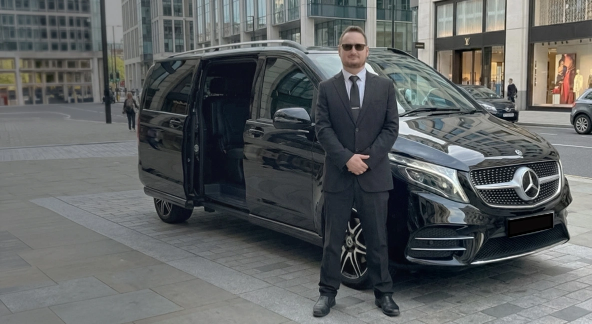 Premium chauffeur-driven Mercedes V-Class for private hire in the UK, suited driver standing beside luxury black MPV in city street
