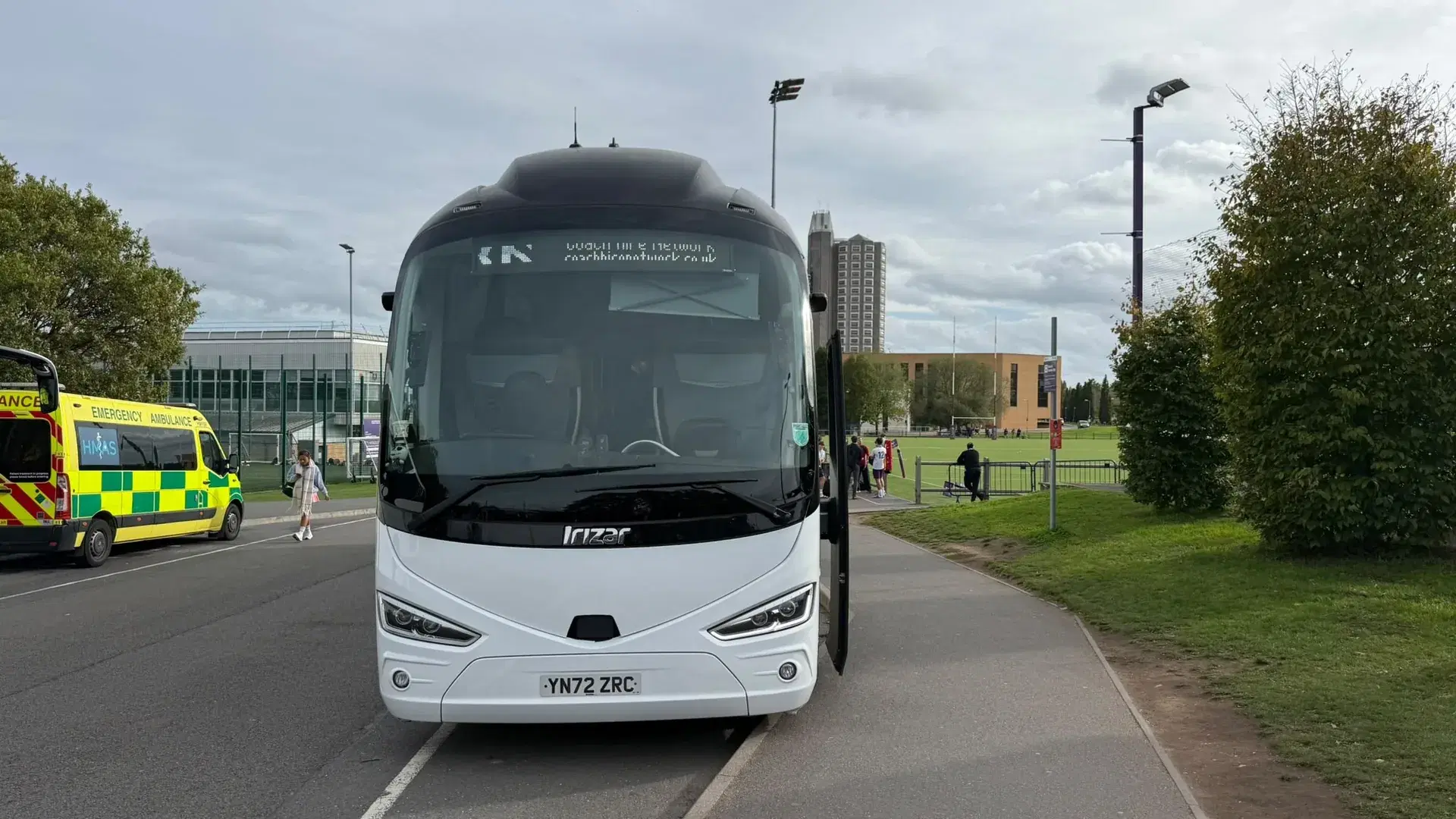 Coach with the door open at a sports pitch, with players on the field behind